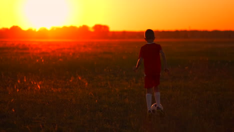 Young-boy-football-player-in-a-red-t-shirt-running-across-the-field-with-the-ball-at-sunset-to-the-sun-camera-on-steadicam